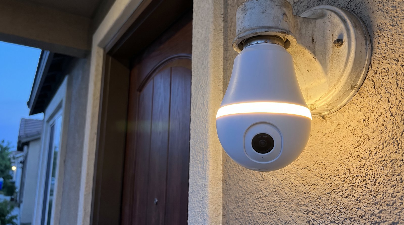 Bulb-shaped security camera with a lens and lit LED ring screwed into a porch light socket at dusk